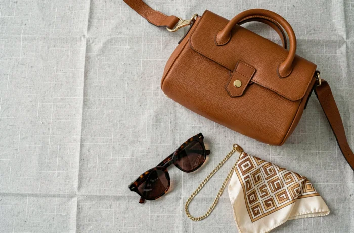 Flat lay d'accessoires de mode incluant un sac en cuir cognac, des lunettes écaille, un carré de soie et un bracelet doré sur lin gris